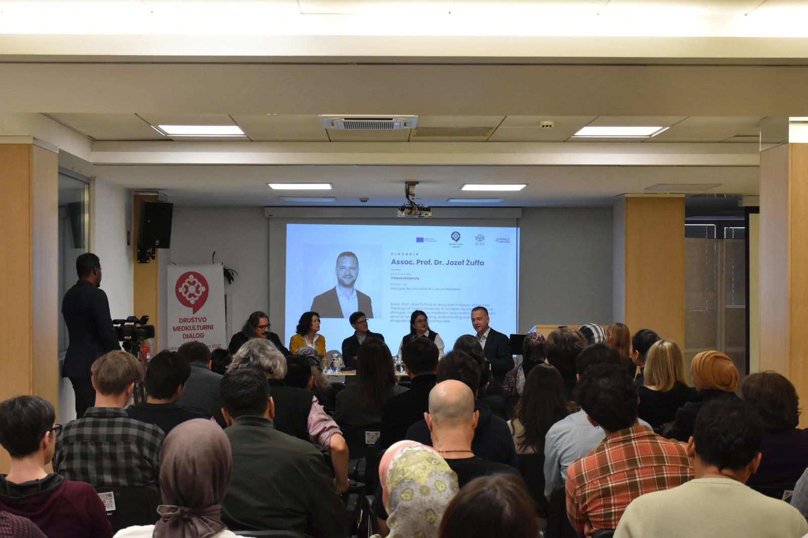 A full hall during the discussion, with a speaker’s profile on the screen